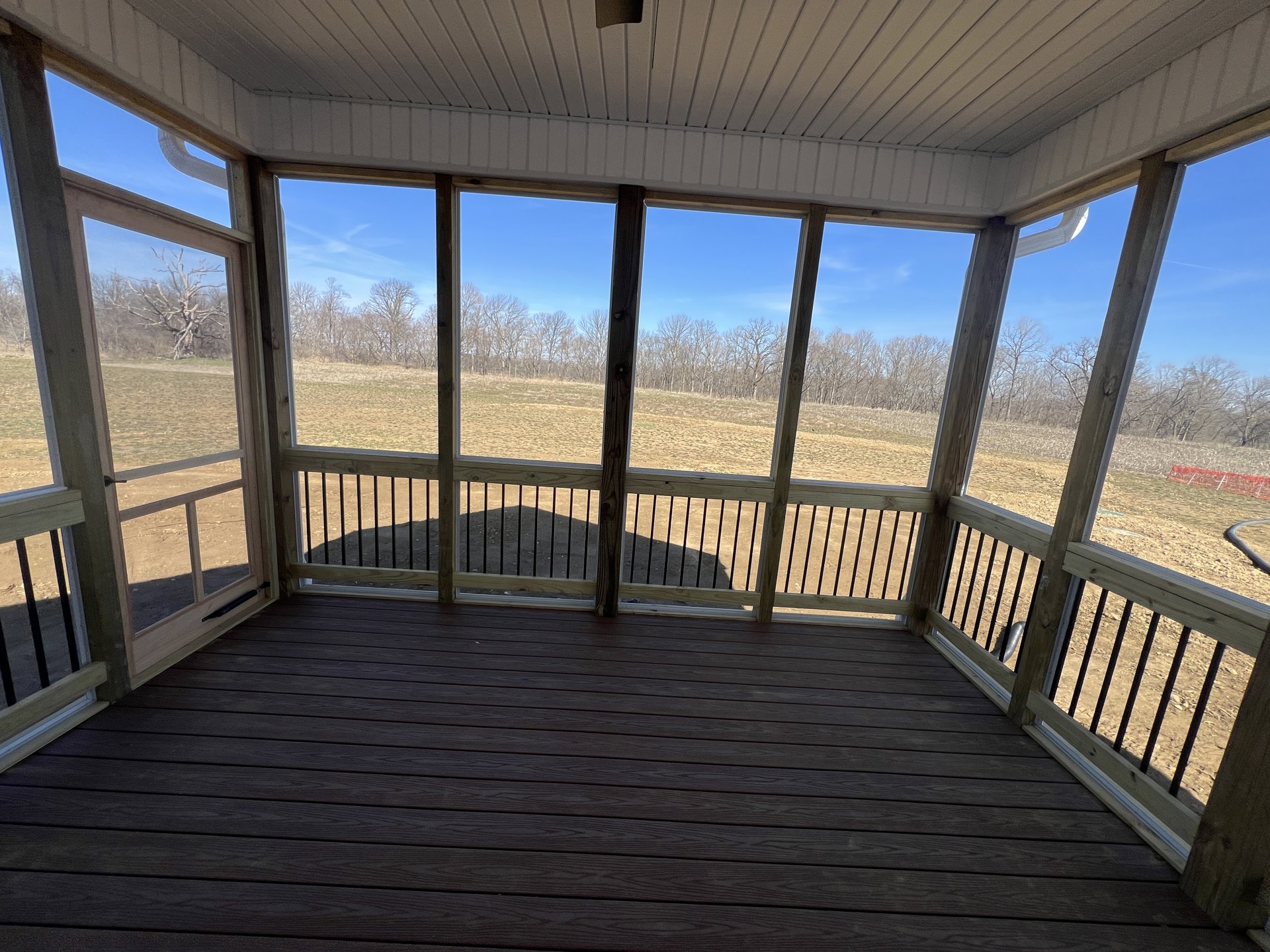 Wooden porch with railing overlooking open field and trees, deck flooring and ceiling visible, window with bars on side wall