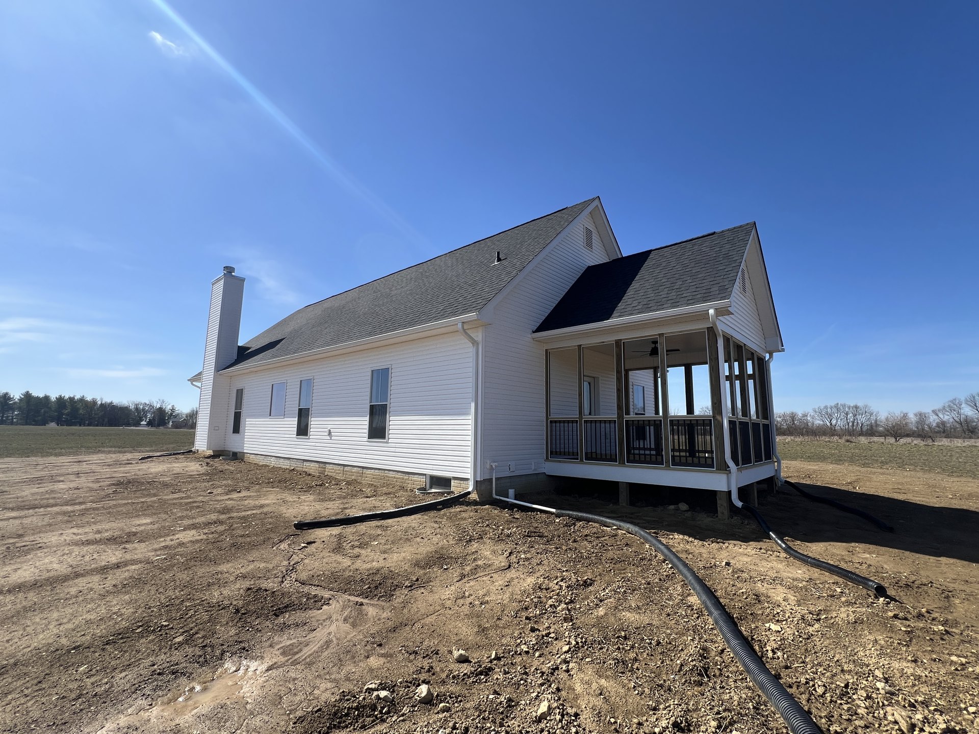White house with black roof and porch under construction, dirt lot with exposed pipe in foreground, large grassy field and group of distant trees, white chimney visible against