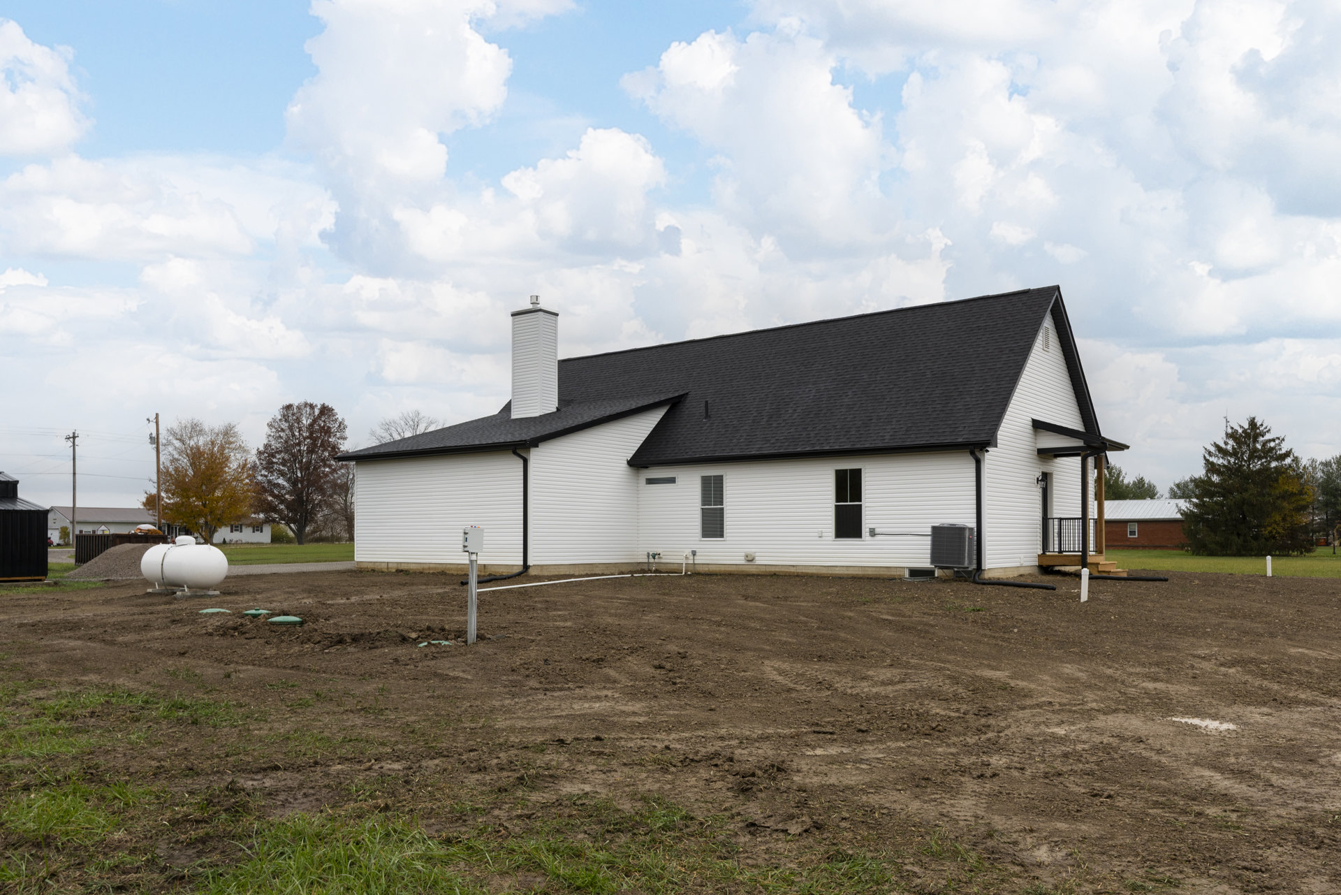 White house with black roof and chimney, large grassy yard, white garage, elevated white tank, tree in front, rural landscape under cloudy sky