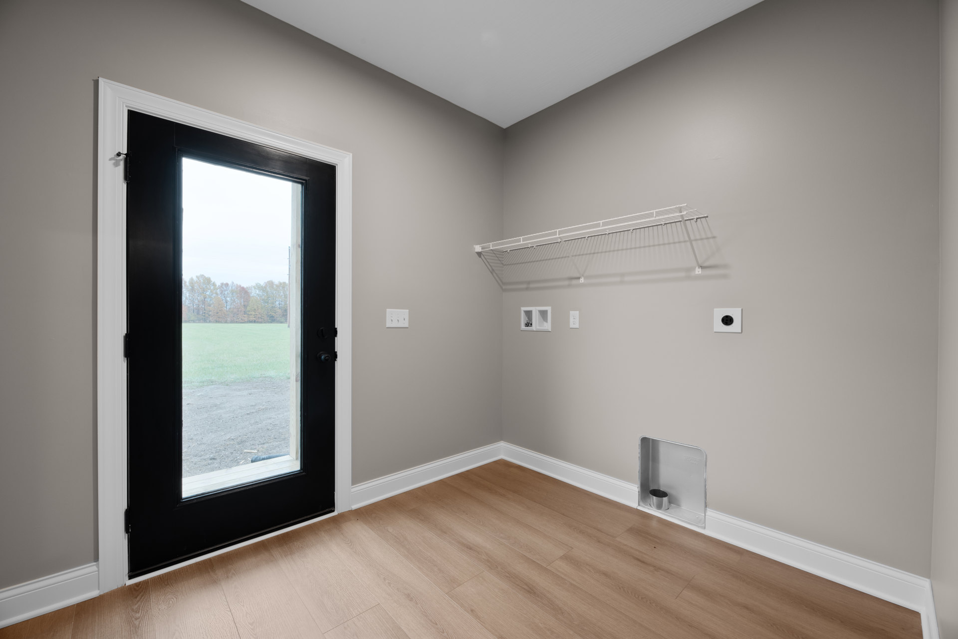 Room with light wood flooring, white baseboards, black door, glass door overlooking field and trees, built-in metal container, and white wall shelf