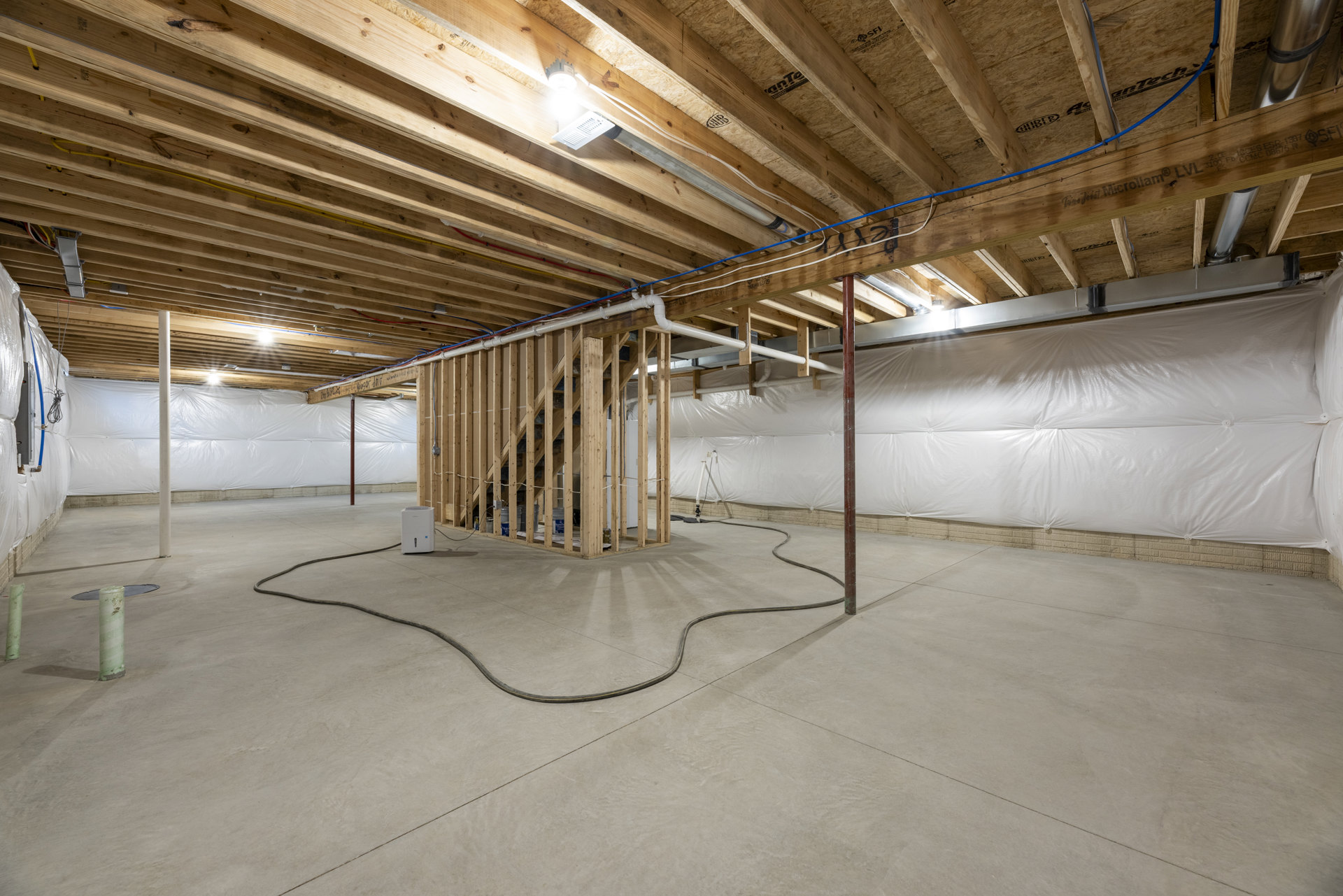 Basement room with exposed wood framing, white sheet covering part of wall, ceiling light, long hose on concrete floor, visible pipe, and white rectangular device with clear lid.