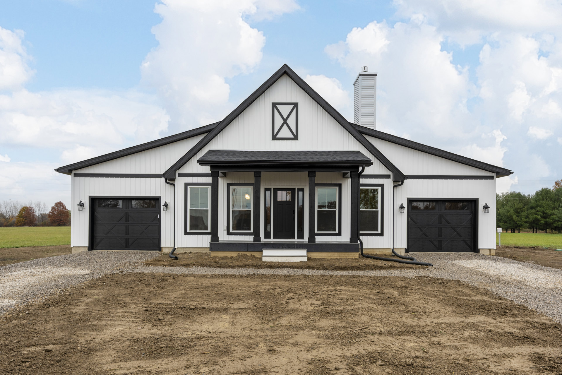 White modern house with black trim, black front door and windows, black pillars, dirt driveway, sparse landscaping, cloudy sky.