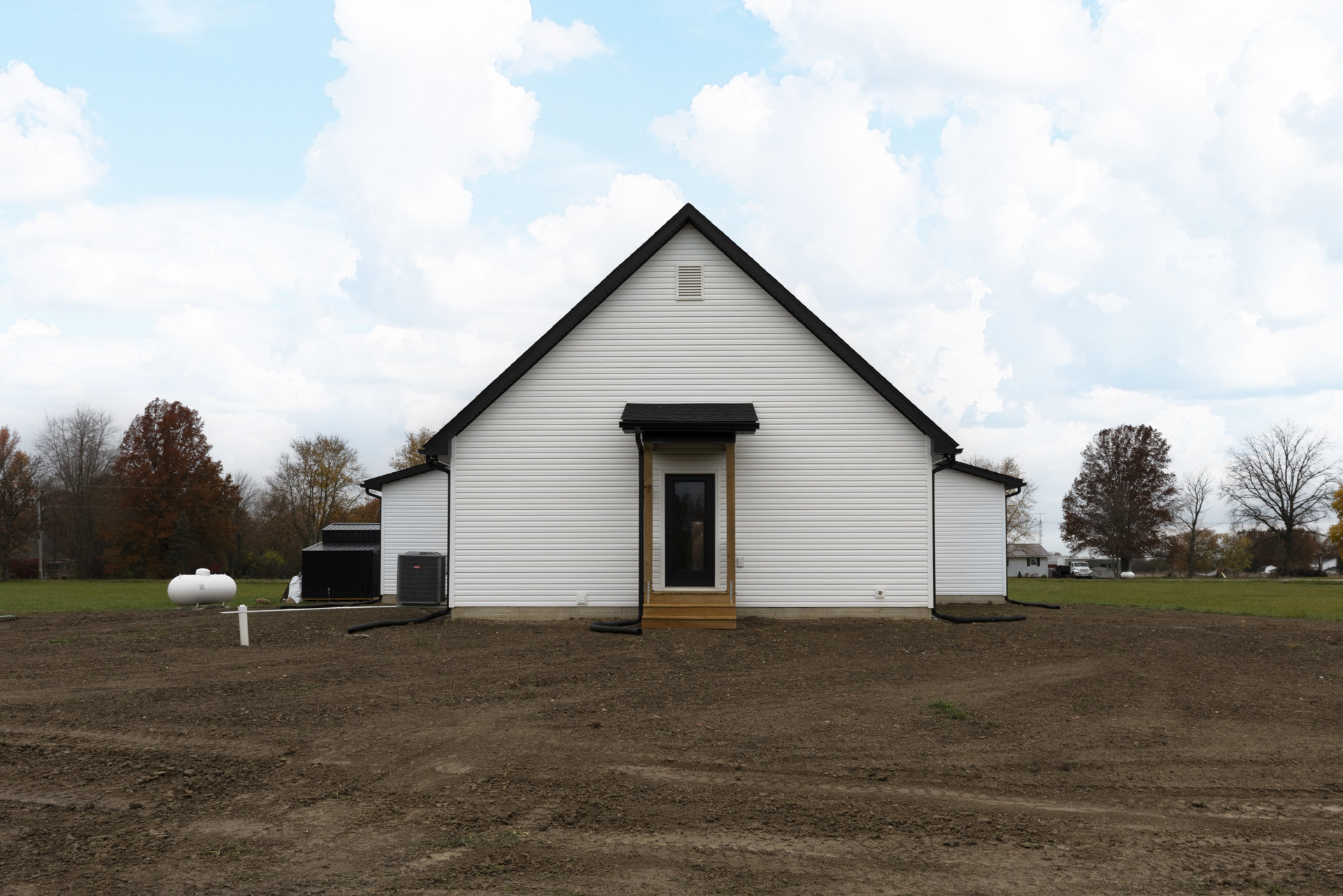 White siding farmhouse with black shingle roof, black front door, leafless tree, white water tank on metal stand, grassy yard, cloudy sky