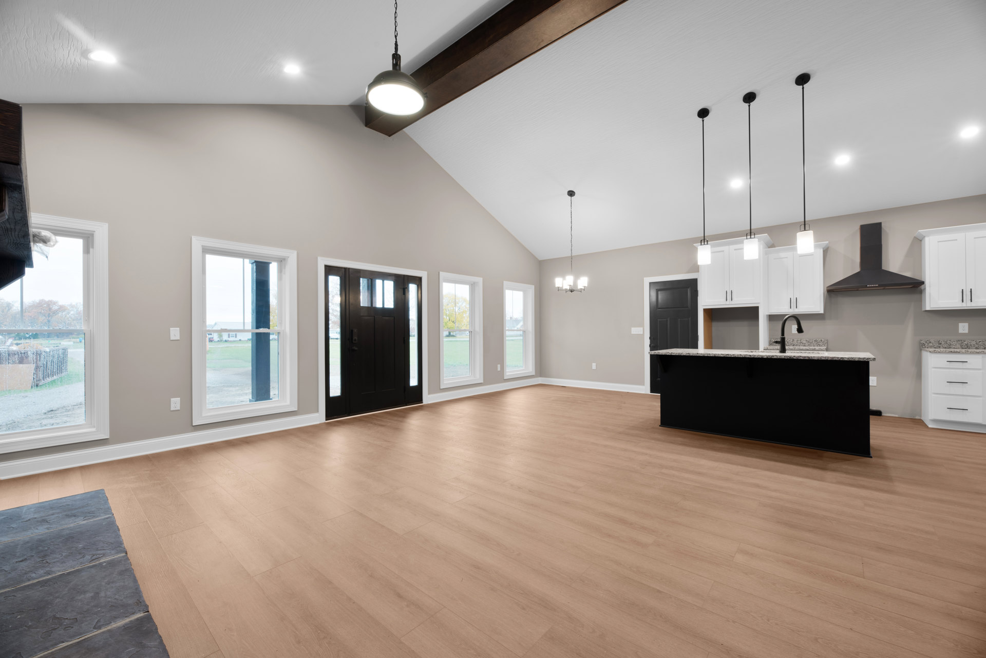 Open-concept kitchen with wood flooring, black cabinetry and countertops, black door with glass panels, large window overlooking grassy field, and grey rectangular tile inset in