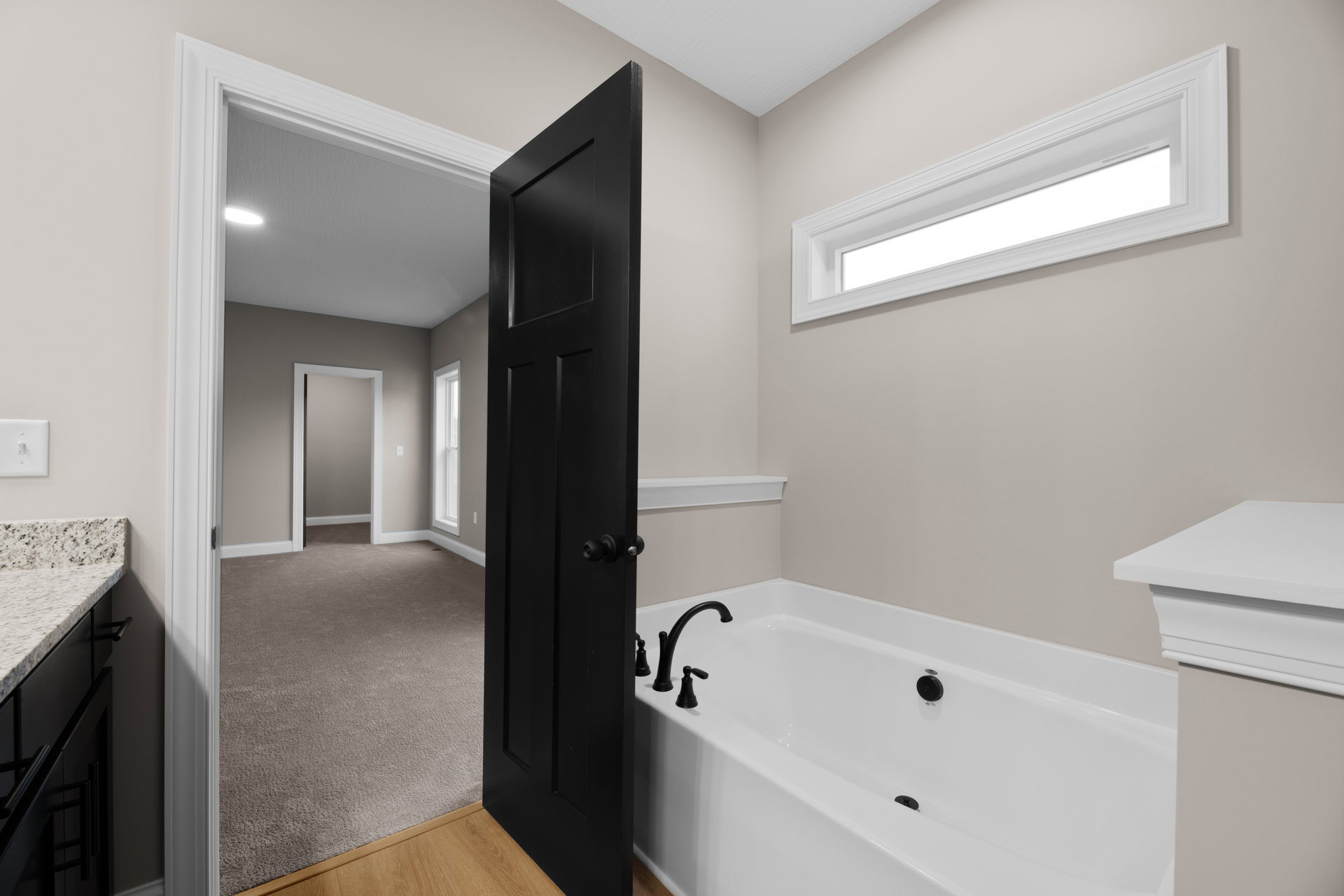 White bathtub with black drain beside a black door with round knob, white ceiling with recessed light, grey walls, white door frame, and carpeted floor.