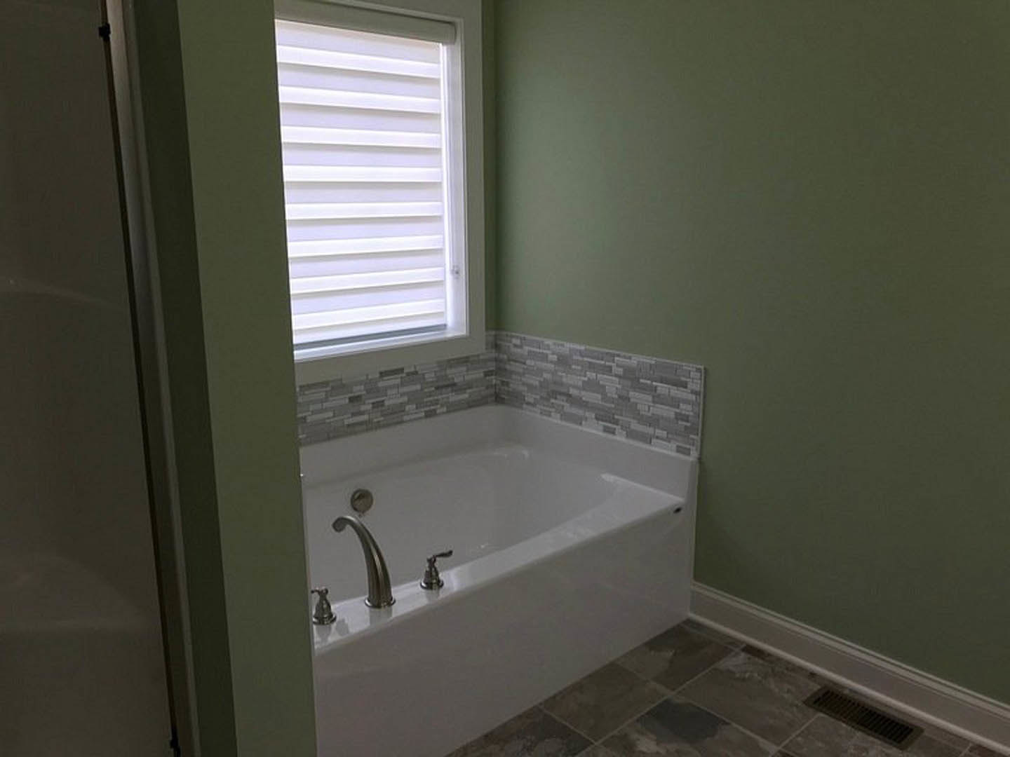 Freestanding bathtub with black metal faucet beside a window with white blinds, tile floor featuring a floor vent, partial view of bathroom door