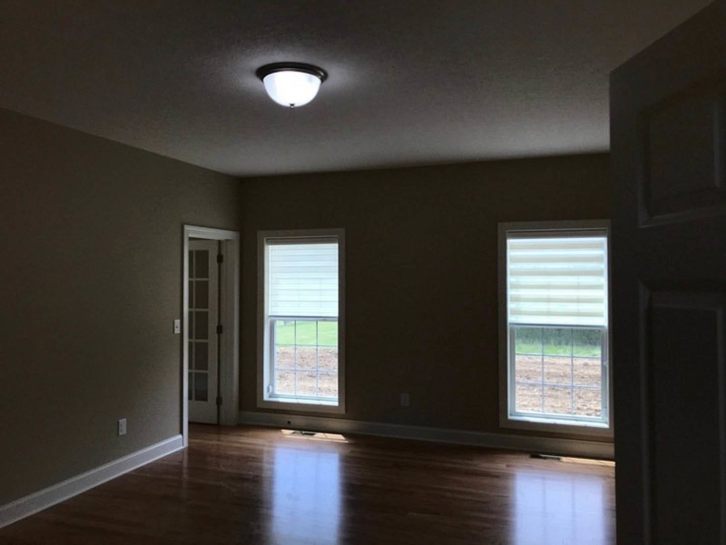 Hardwood floor room with white walls, ceiling light fixture, door with modern trim, two windows—one with white shade, one overlooking green field