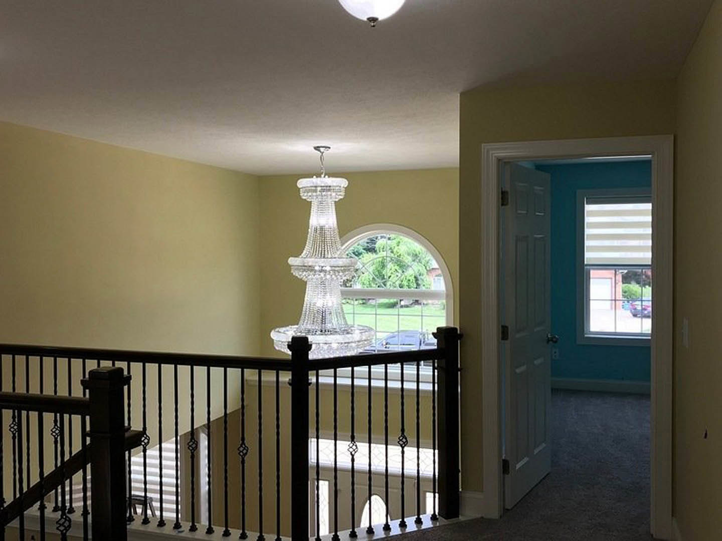 Wrought iron staircase with wood handrail, ornate chandelier hanging above, white and brown striped window shade, open door revealing window view of green lawn