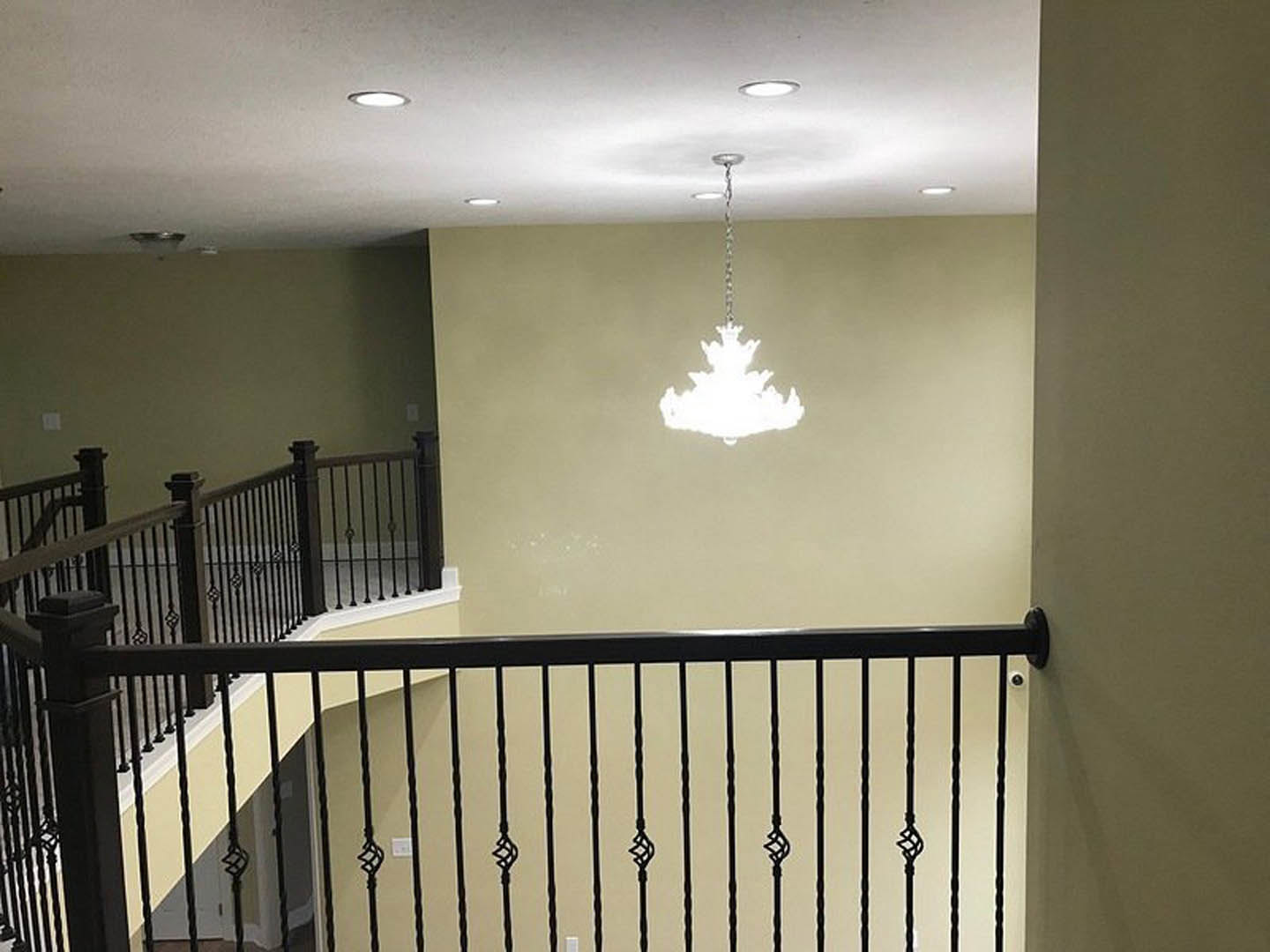 Black iron staircase railing against white plaster wall, modern chandelier hanging from ceiling, recessed lighting illuminating entryway.