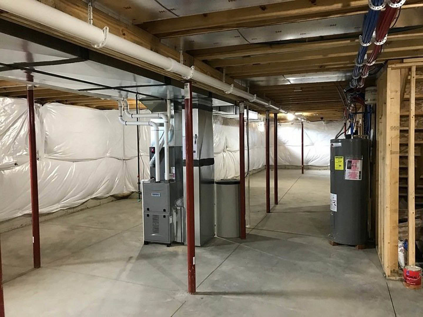Basement utility room with exposed steel and aluminum pipes along the ceiling, grey cylinder and box mounted on a white wall with black border, large white bag attached to wall