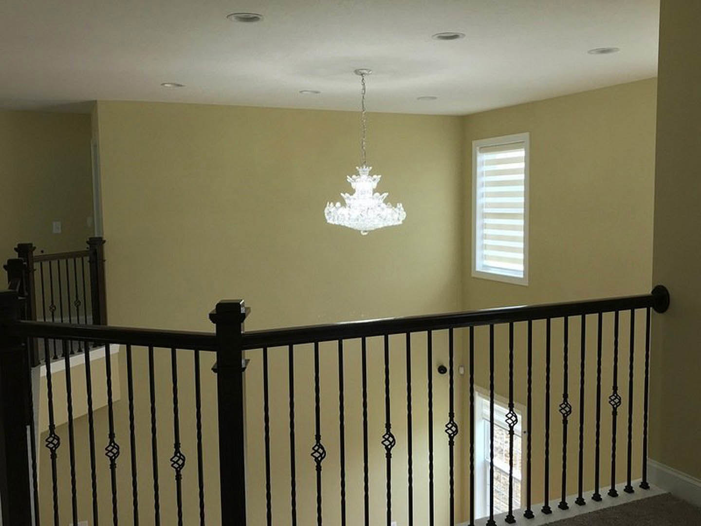 Chandelier with glass accents suspended from white ceiling, black iron railing along staircase, white plaster walls, window with light fixture visible, decorative molding details