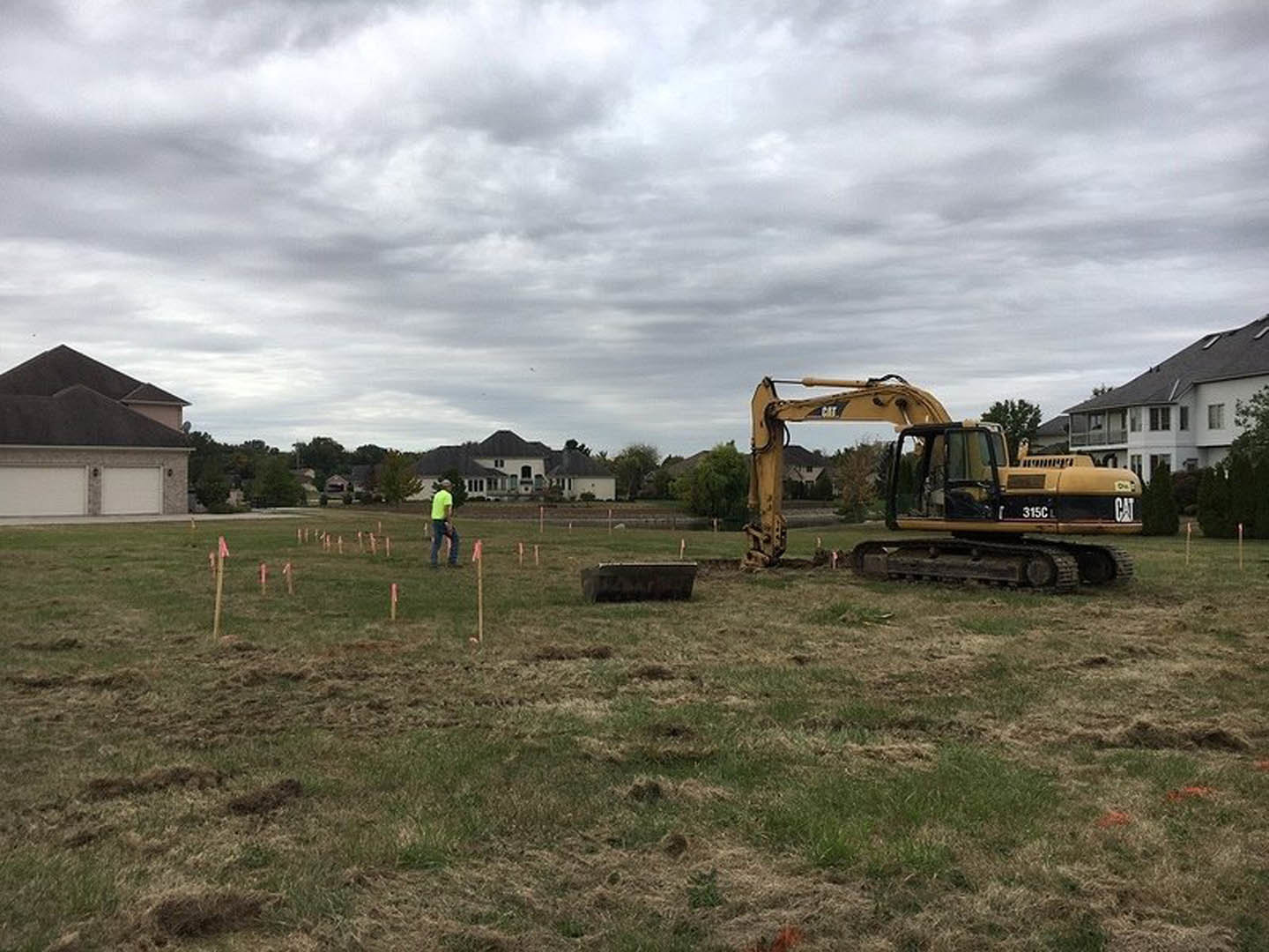 Bulldozer parked on grassy lot beside partially built custom home, cloudy sky overhead, trees and construction materials in background