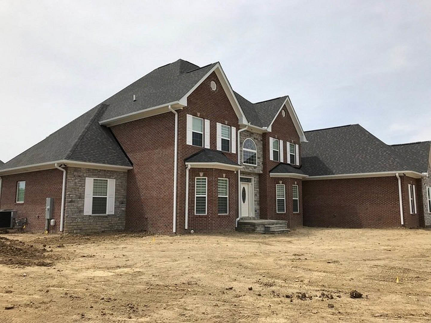Brick house with white door and white-framed windows, white shutters, dirt yard in foreground, cloudy sky overhead