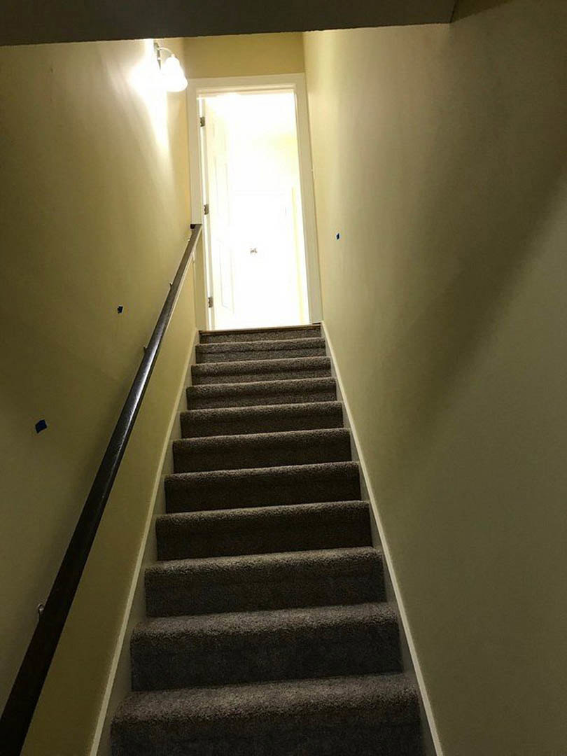 Wood staircase with metal handrail ascending toward a white door, bright natural light streaming from above, smooth white walls and light hardwood flooring