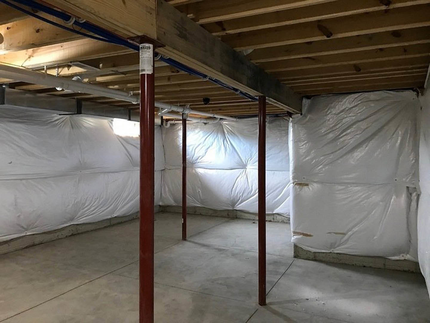 Basement with white plastic wall coverings, exposed wooden ceiling beams, concrete floor, and structural pillar