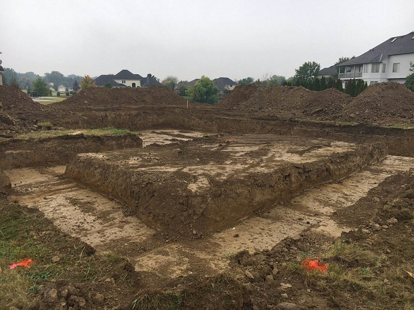Expansive dirt lot with scattered soil piles, bordered by green grass and trees, custom home with balcony visible in background under blue sky