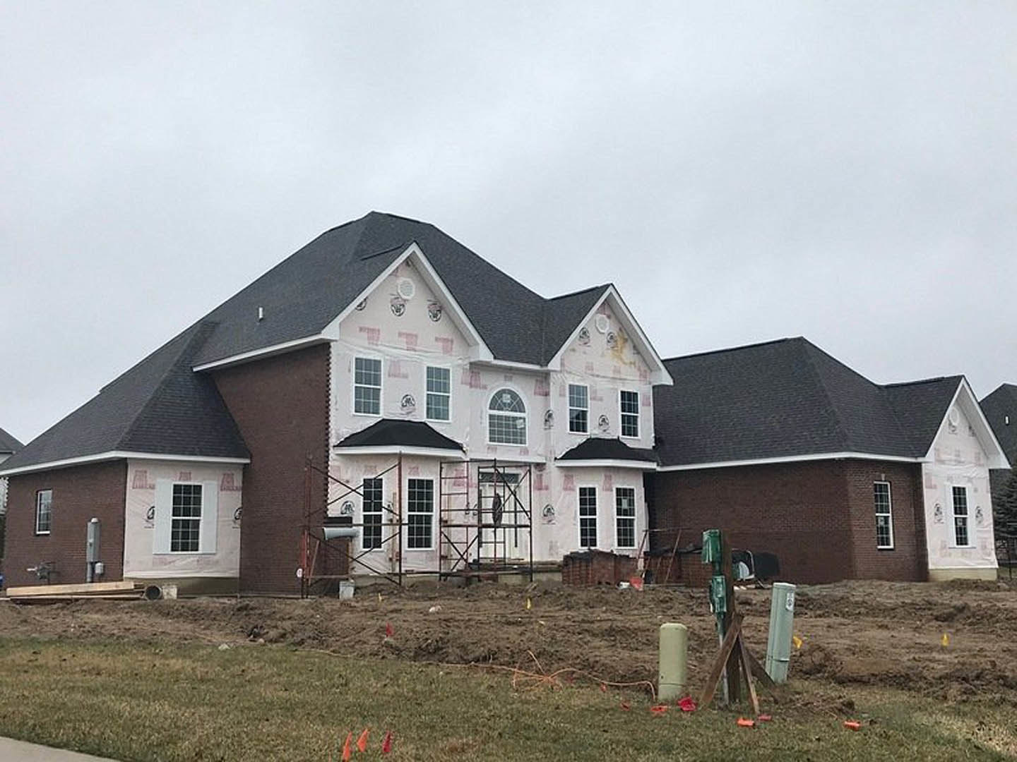 Scaffolding surrounds a partially built house with a black roof, square-paned windows, and exposed framing; white posts and a grassy lawn are visible in the foreground.