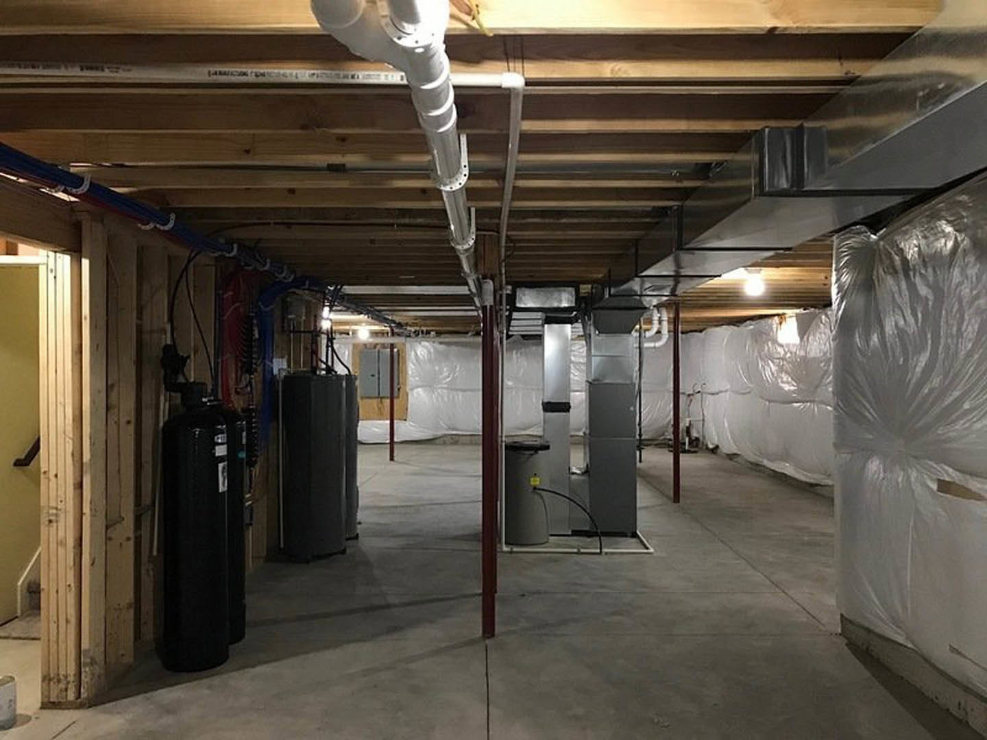 Unfinished basement room with exposed white pipes along the ceiling, yellow utility box mounted on wall, black rectangular object on floor, plastic wrap covering construction