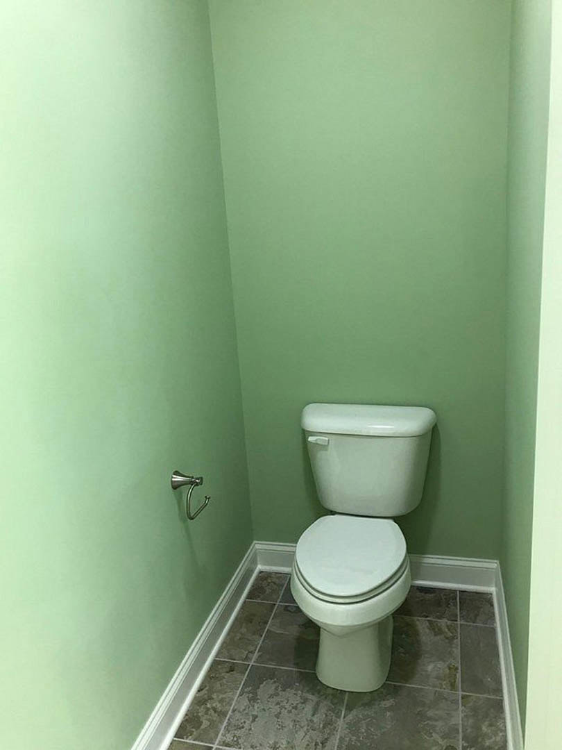 White toilet with lid up against green tiled wall, chrome flush handle on tank, silver bathroom accessory mounted nearby