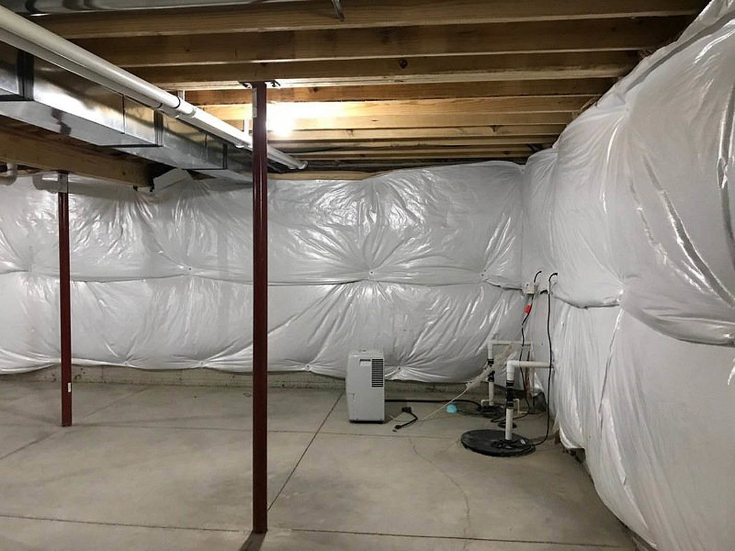 Basement with white plastic coverings on walls and floor, exposed steel support pole, white rectangular vent, plastic bag attached to wall, microphone resting on table