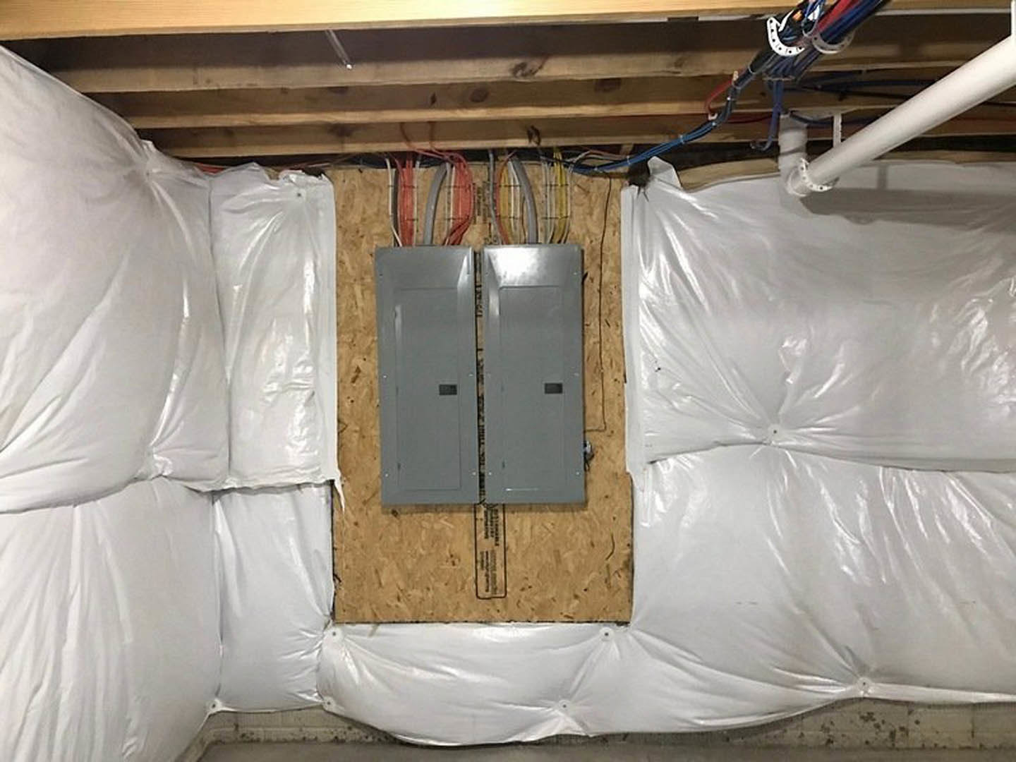 Wall covered in clear plastic sheeting with exposed electrical boxes and wires, grey door, wooden shelf, white plastic bag, and visible building insulation in a room under remodel.