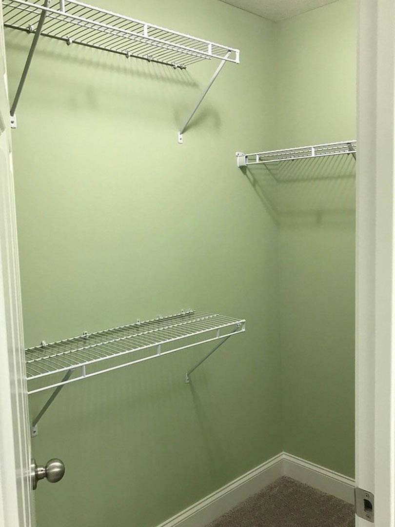 Walk-in closet featuring white shelving units, metal hanging rods, light green painted wall, beige carpet flooring, and a white door with a brushed metal knob