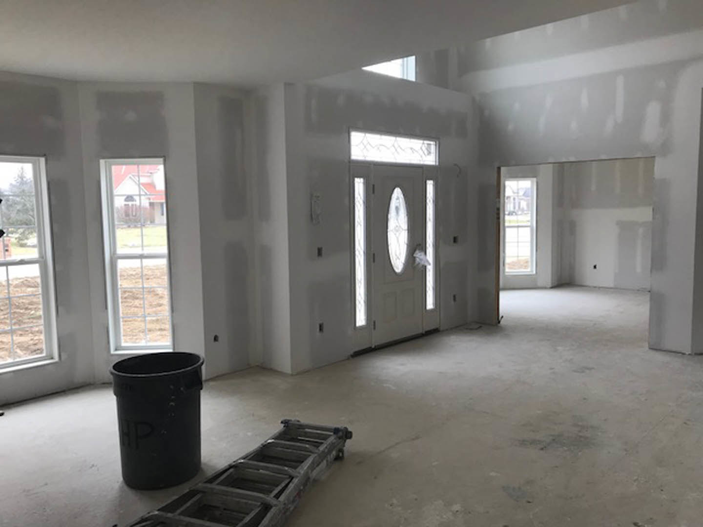 White paneled door with frosted glass window, black trash can with handle beside wooden ladder, plaster walls, light wood flooring, window overlooking grassy field