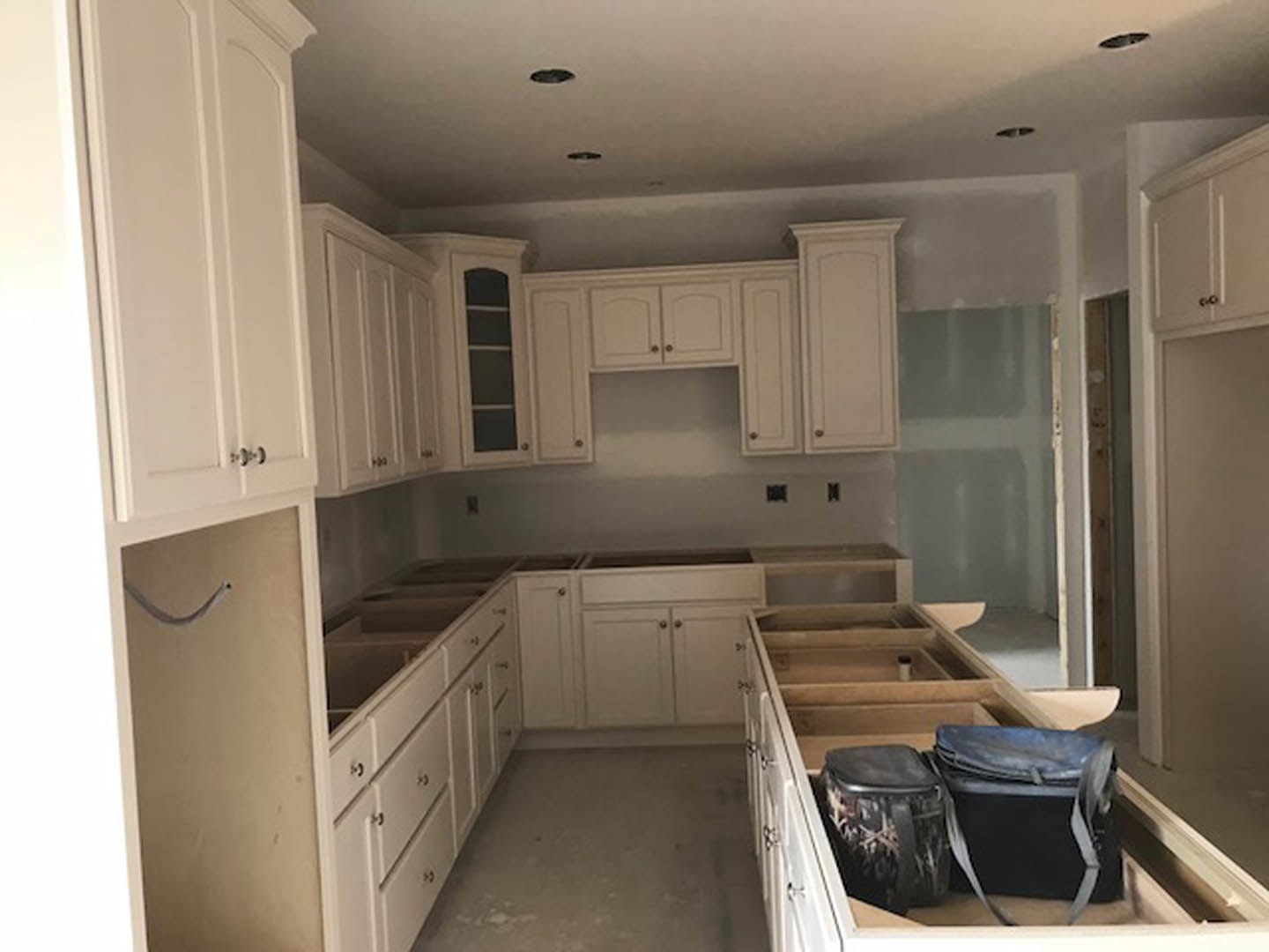 White kitchen cabinets, wood flooring, several bags near the island, stainless steel appliances, white-framed window above the sink