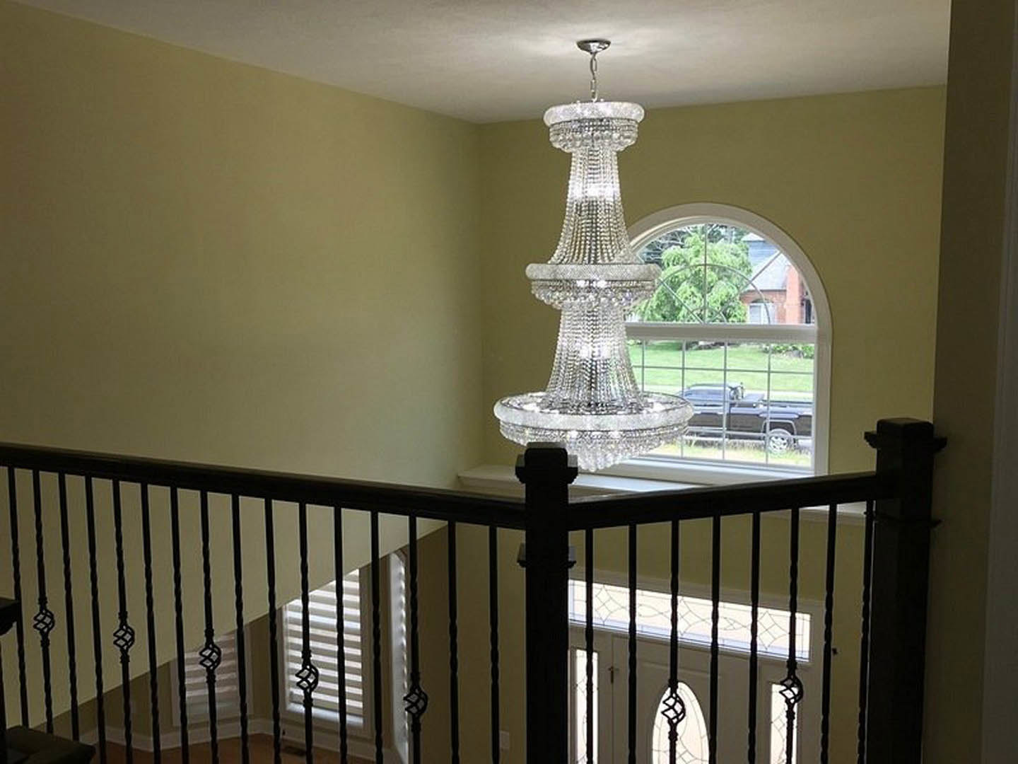 Crystal chandelier hanging above staircase with iron balusters, white ceiling, and window showing black truck parked behind fence outside.