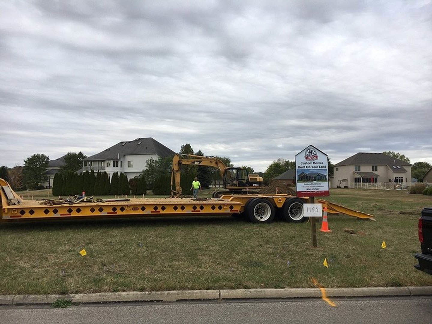 Yellow flatbed trailer parked on grassy field beside fenced custom home, white sign with black numbers and house illustration in foreground, cloudy sky overhead.