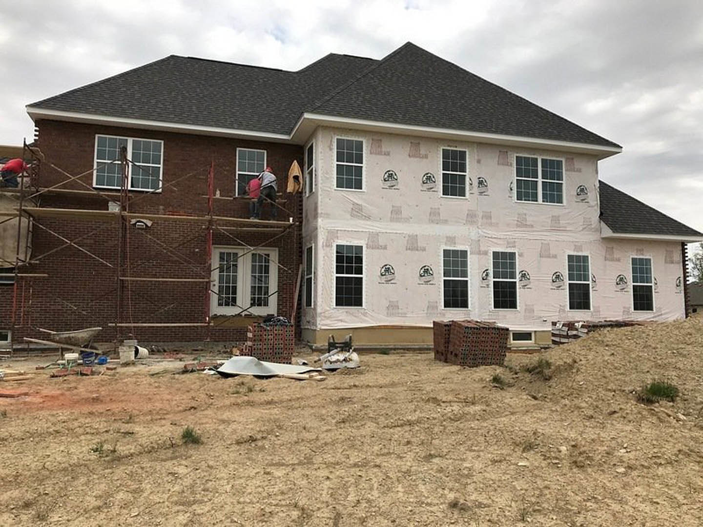 Two workers on scaffolding installing exterior finishes on a partially built house, with stacks of bricks and construction materials on a dirt lot, windows visible on the