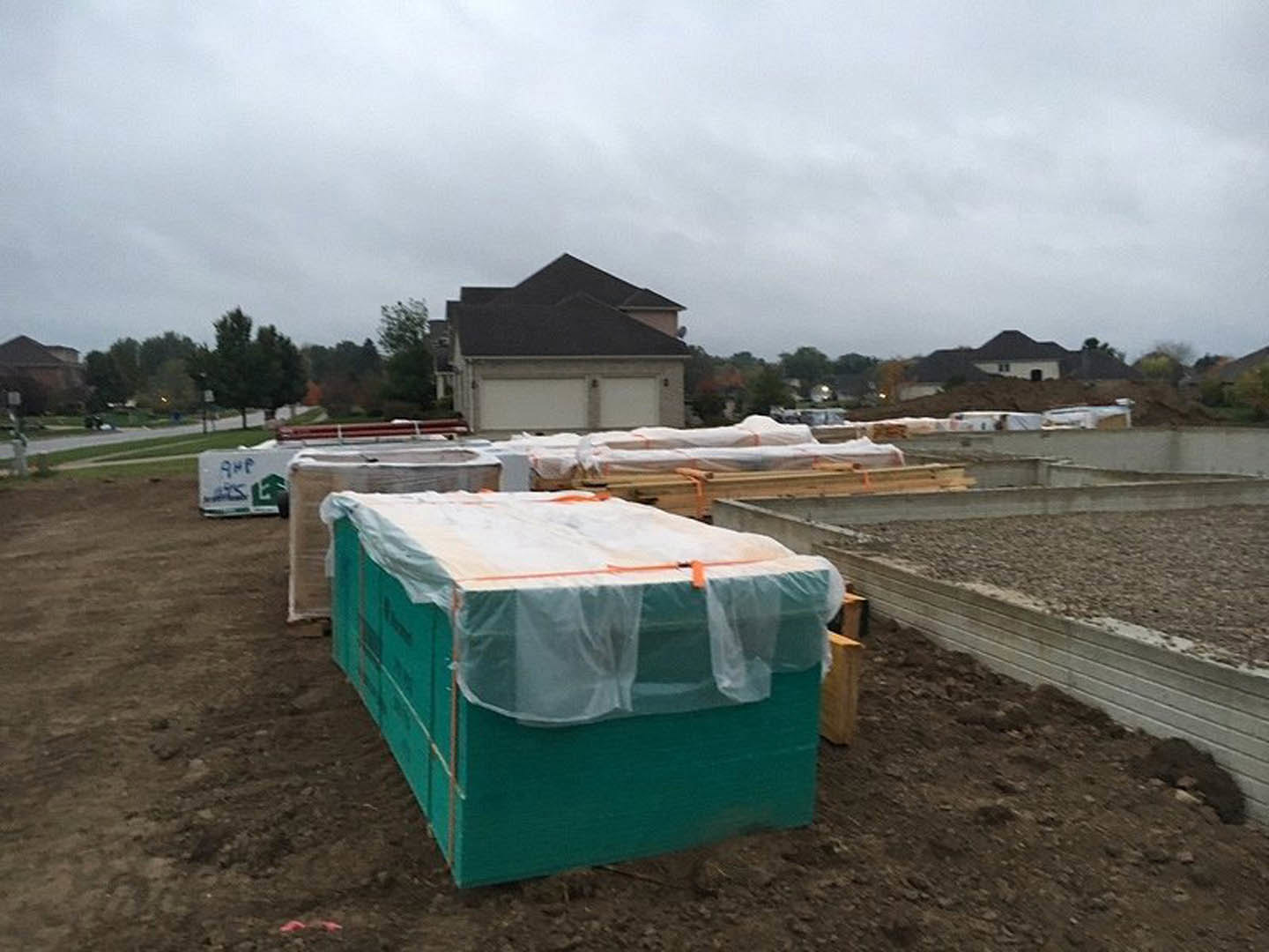 Partially built house with exposed roof, plastic-covered green utility box, dirt ground, and cloudy sky