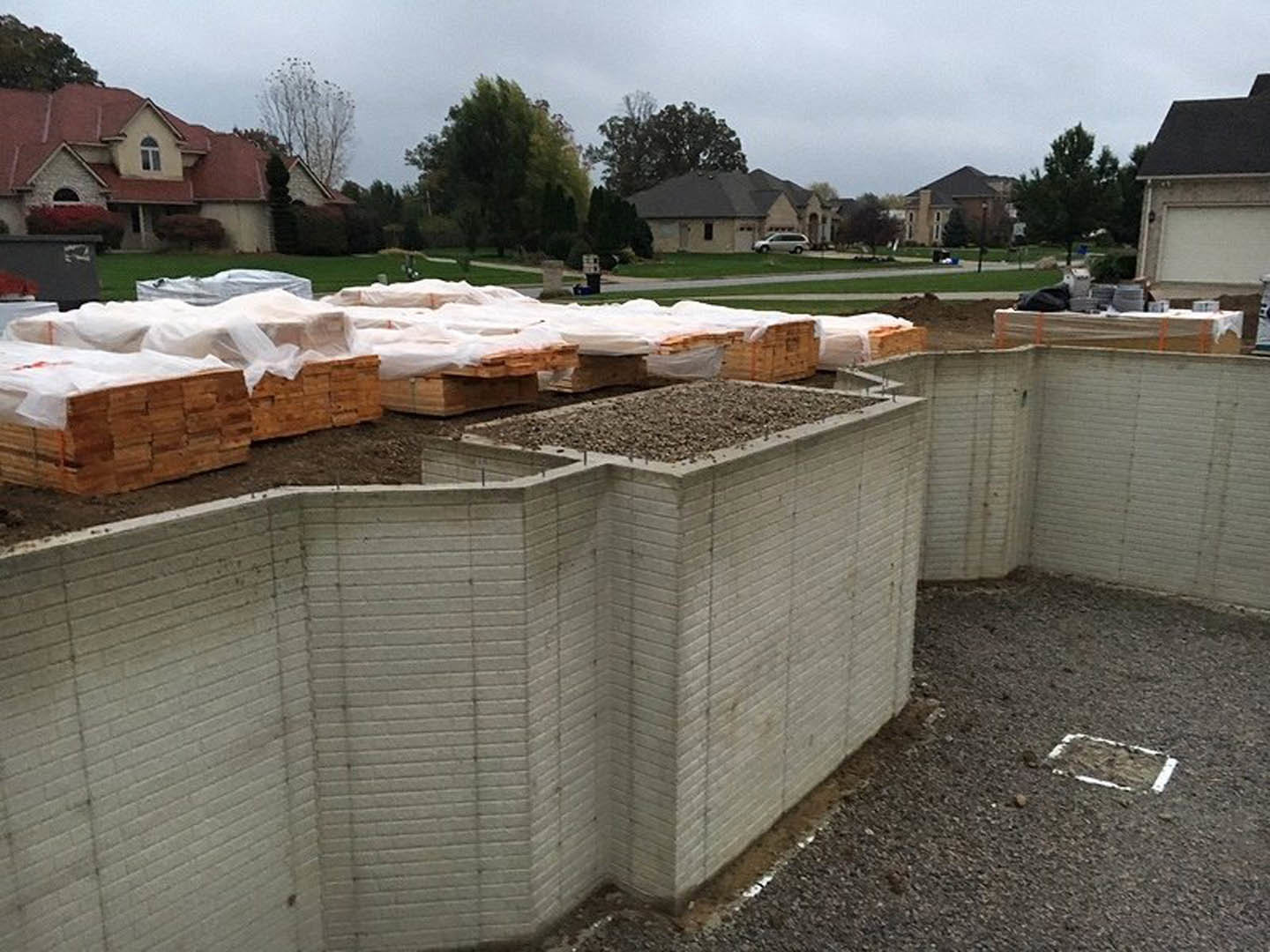 Red-roofed house beside a concrete wall, gravel ground with stacked pallets, large leafy tree, wooden box with white sheets, white line marked in dirt.