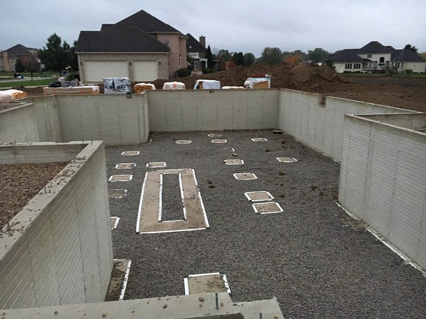 Concrete foundation and framing surrounded by dirt and gravel, partially constructed custom home with black roof, outdoor setting under cloudy sky
