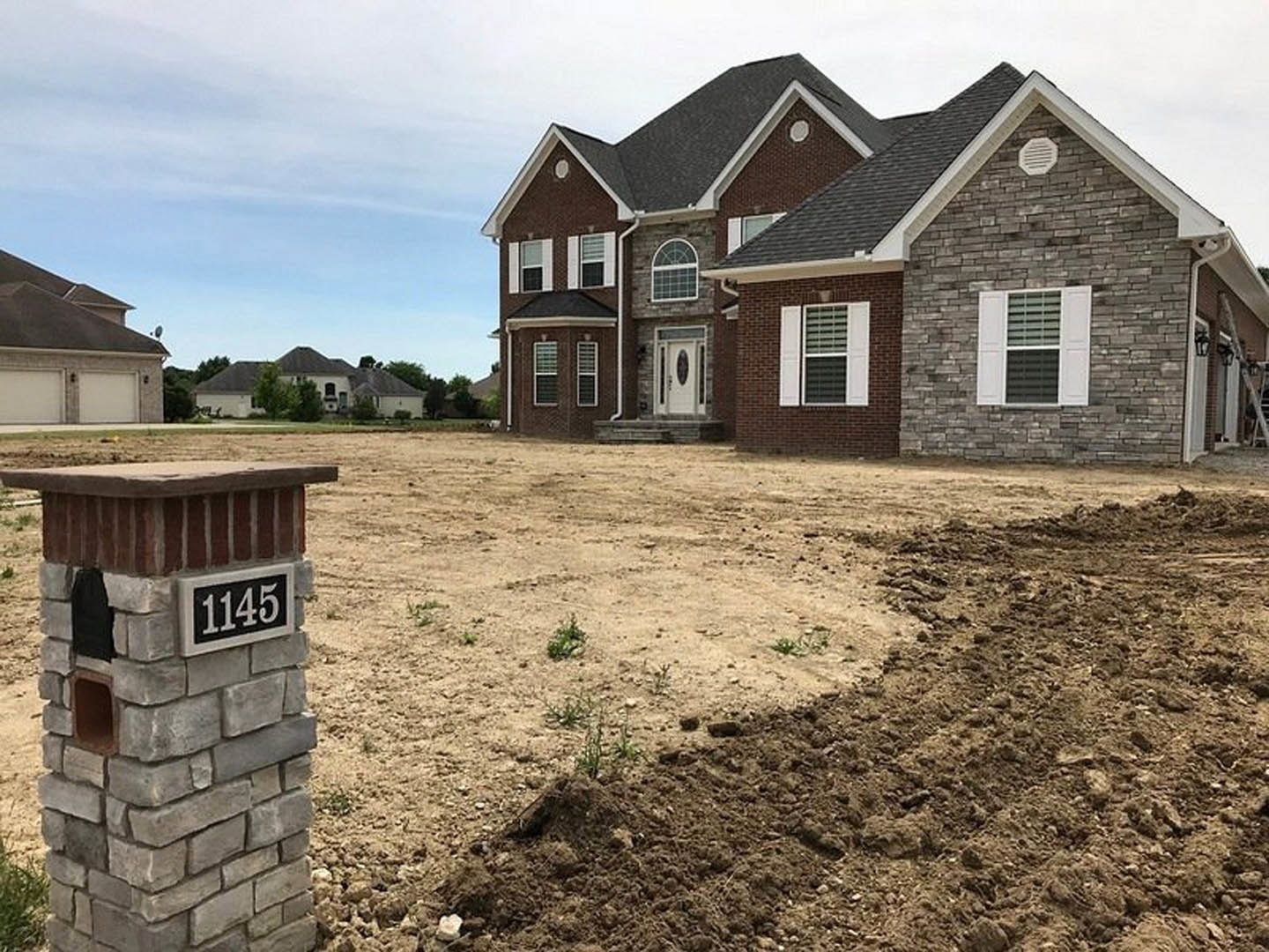 Two-story house with white-framed windows, brick mailbox displaying house number, black and white address sign, brick wall vent, and window with dark shutters, set beside a dirt