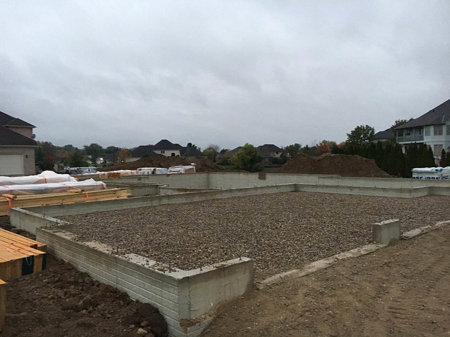Concrete house foundation with dirt, gravel, and scattered rocks under a cloudy sky, stack of wood nearby, trees in the background