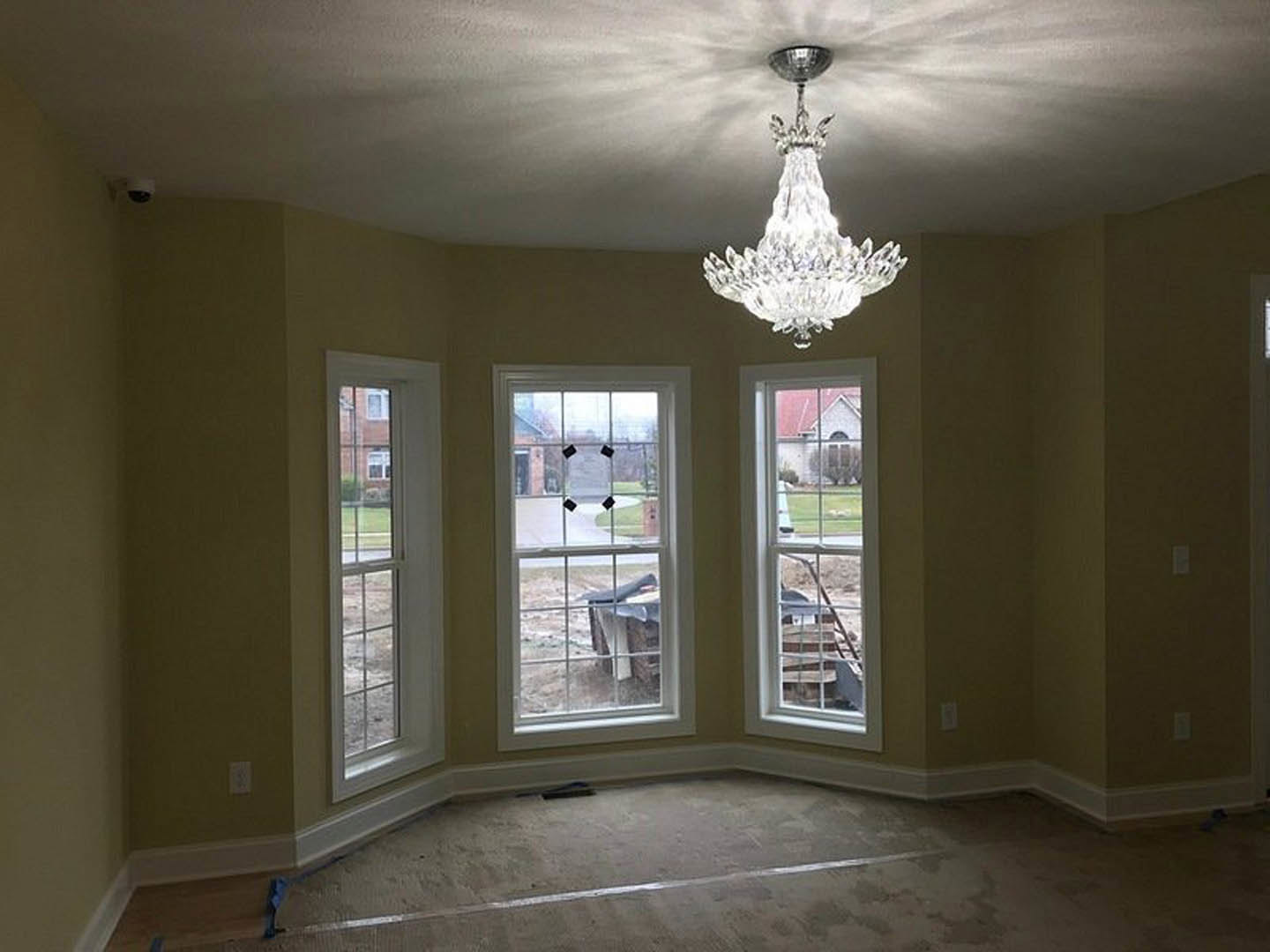 Crystal chandelier hanging from a plaster ceiling in a spacious room with large windows, black window tape, and concrete flooring marked by a white line, overlooking a landscaped
