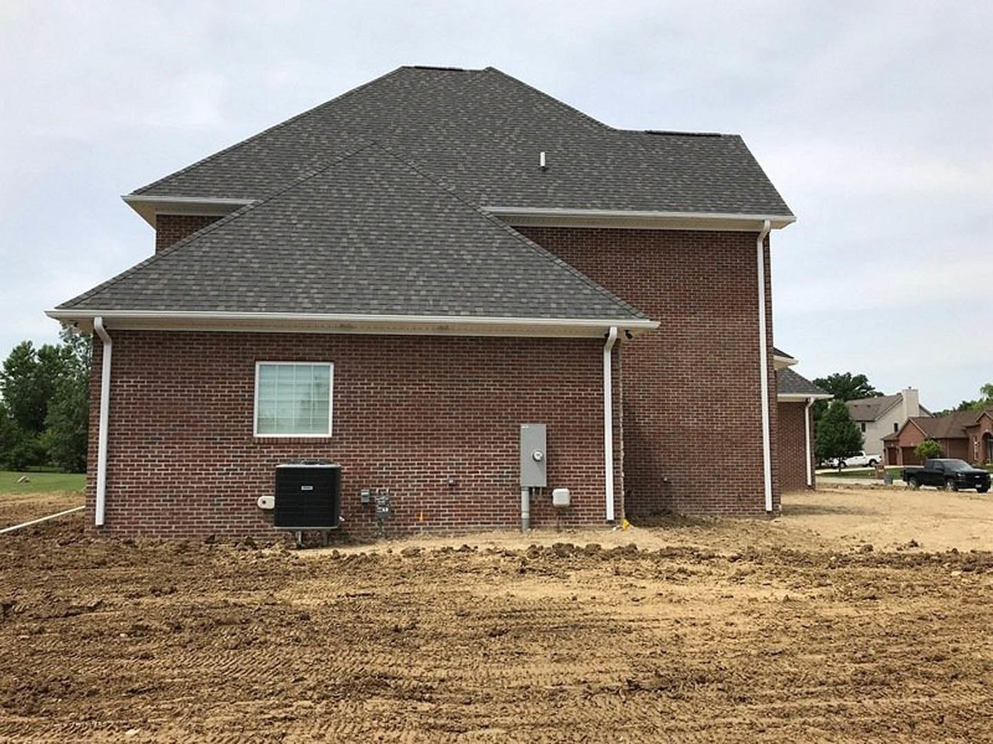 Red brick house with white-framed window, black wall-mounted heater, and large dirt yard; blurred car partially visible, grassy field and trees in background