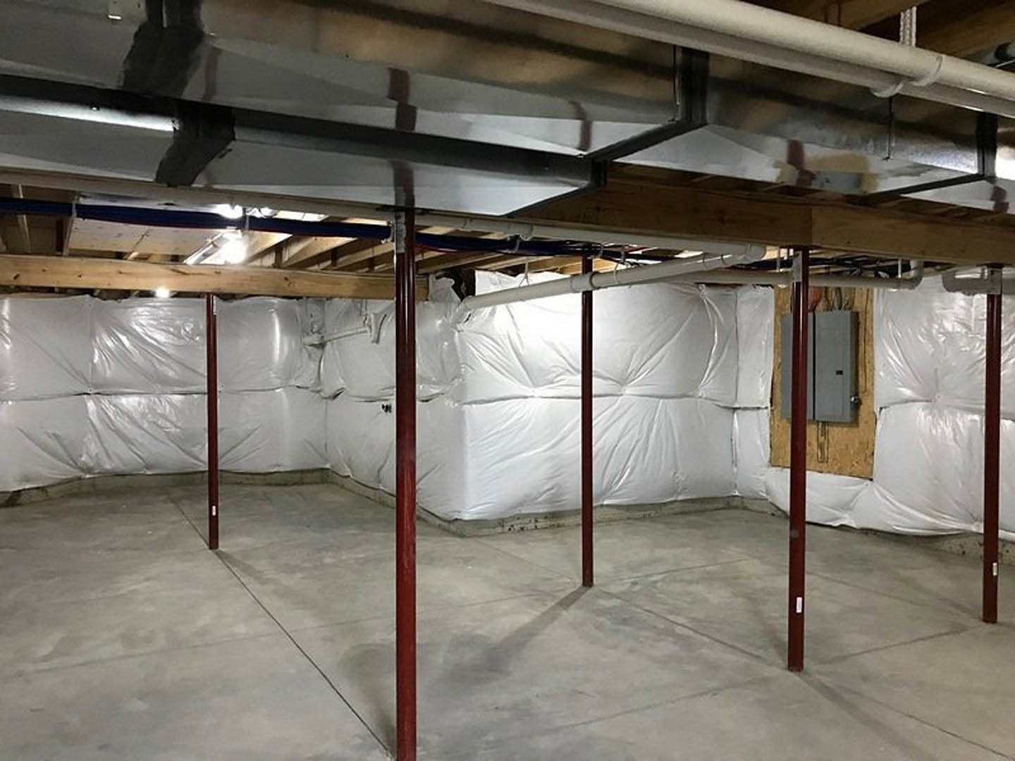 Basement room with exposed beams and ceiling wrapped in white plastic, large white bag on concrete floor, unfinished walls and visible insulation