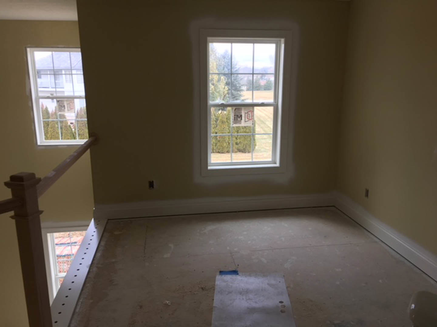 Concrete floor with a piece of paper near a person standing, plaster walls, large window showing neighboring building and house, natural daylight filling the room