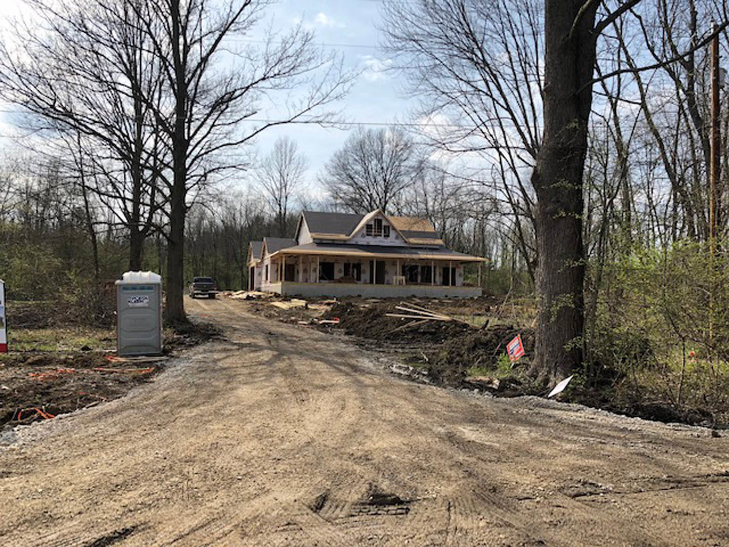 Modern home under construction surrounded by tall trees, grey exterior walls, exposed framing, dirt lot, cloudy sky overhead