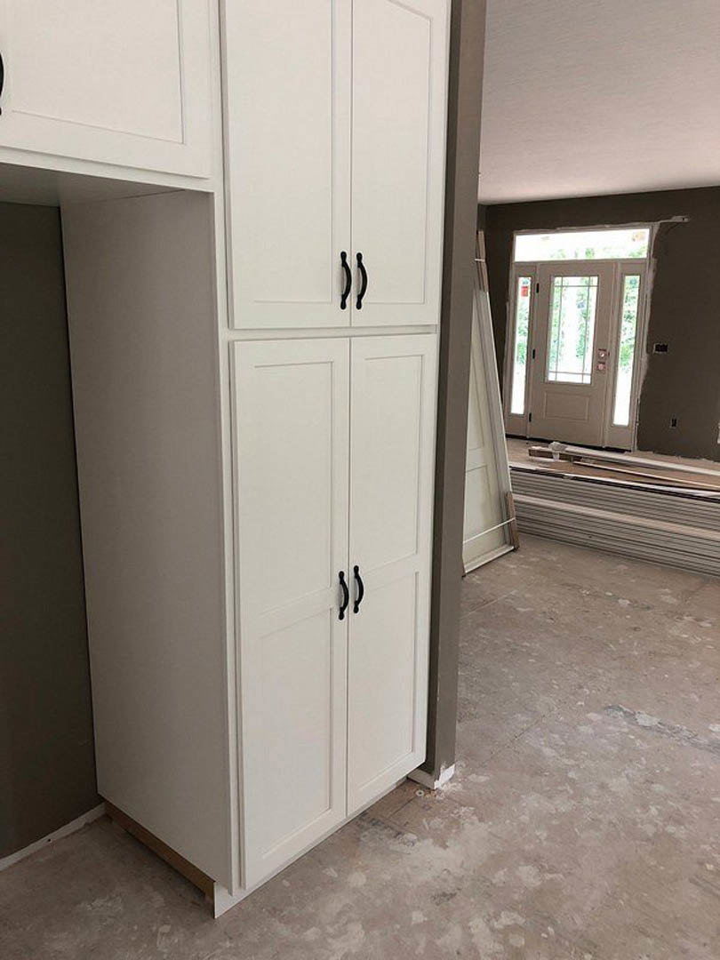White shaker-style cabinet with black handles beside a white glass-paneled door, black accent wall with white trim, white towel hanging from door, stack of metal sheets on floor.