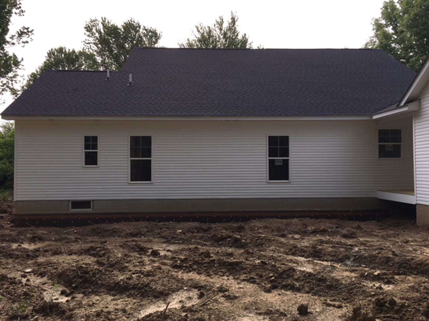 Modern house with black roof, white-framed windows, light siding, and unfinished dirt yard surrounded by trees under a clear sky