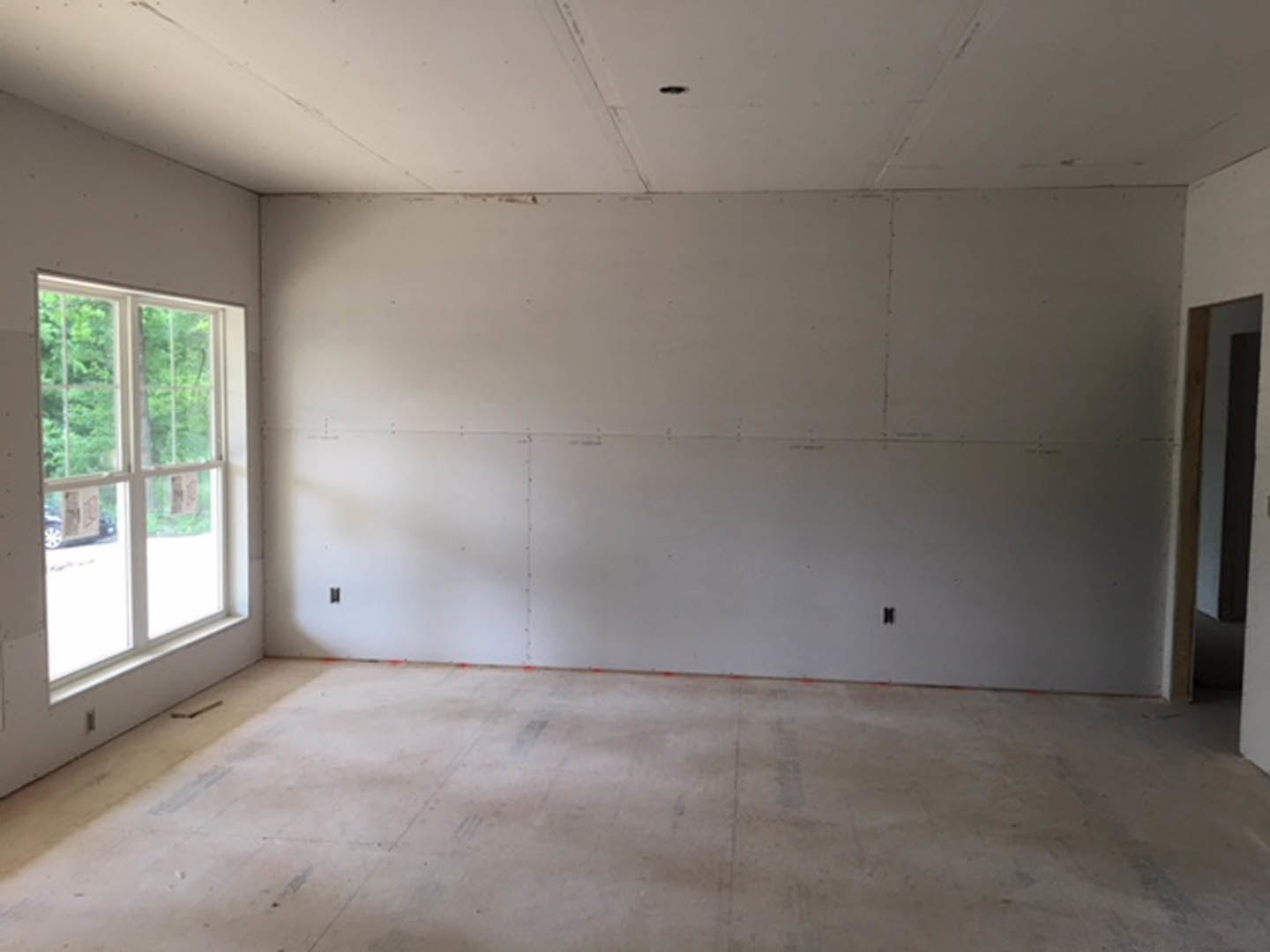 White plaster wall and ceiling, concrete floor marked with red tape, window with papers attached, door partially visible, view of trees outside.