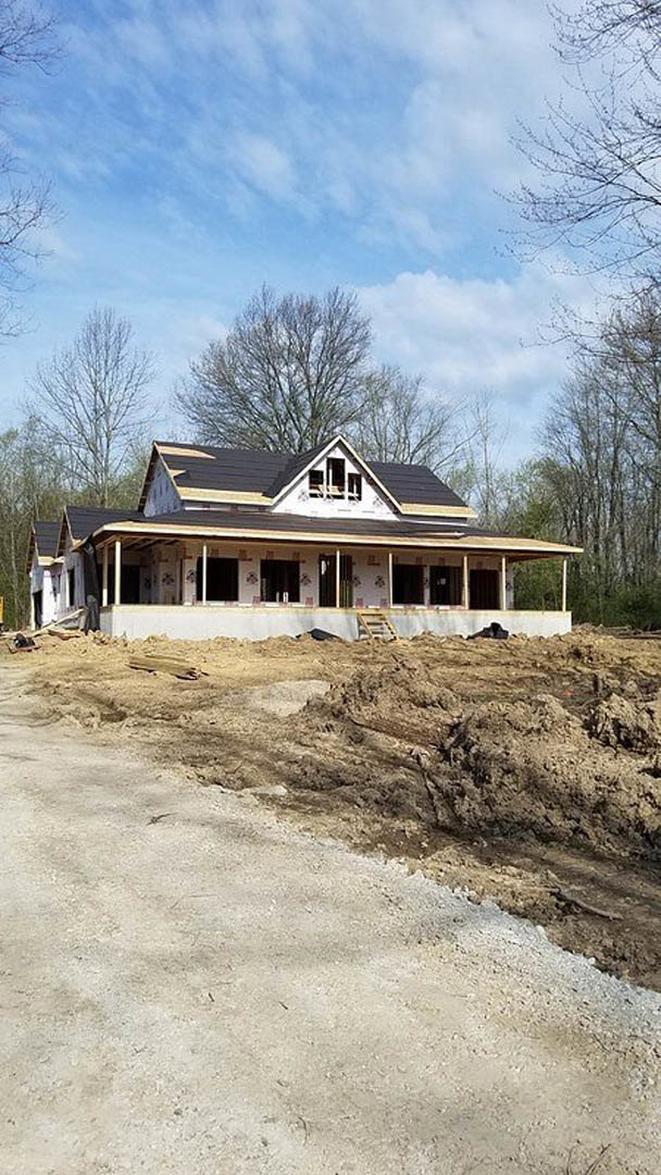Partially built house with black roof and covered porch, surrounded by dirt piles, trees, and blue sky with scattered clouds
