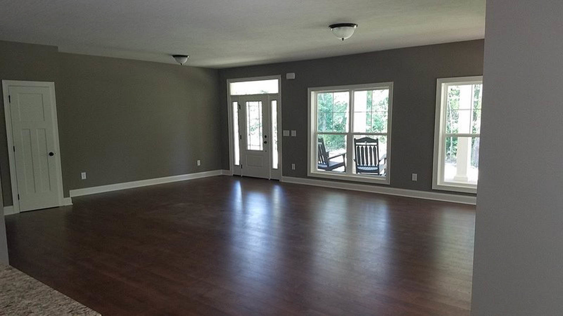 Living room with wide plank wood flooring, large windows, neutral walls, and a modern chair partially visible in the foreground