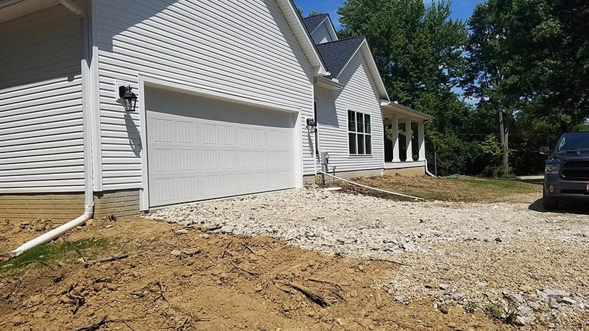 Modern home with attached garage, white siding, dirt patch in front yard, parked car partially visible, leafy trees in background, clear sky overhead