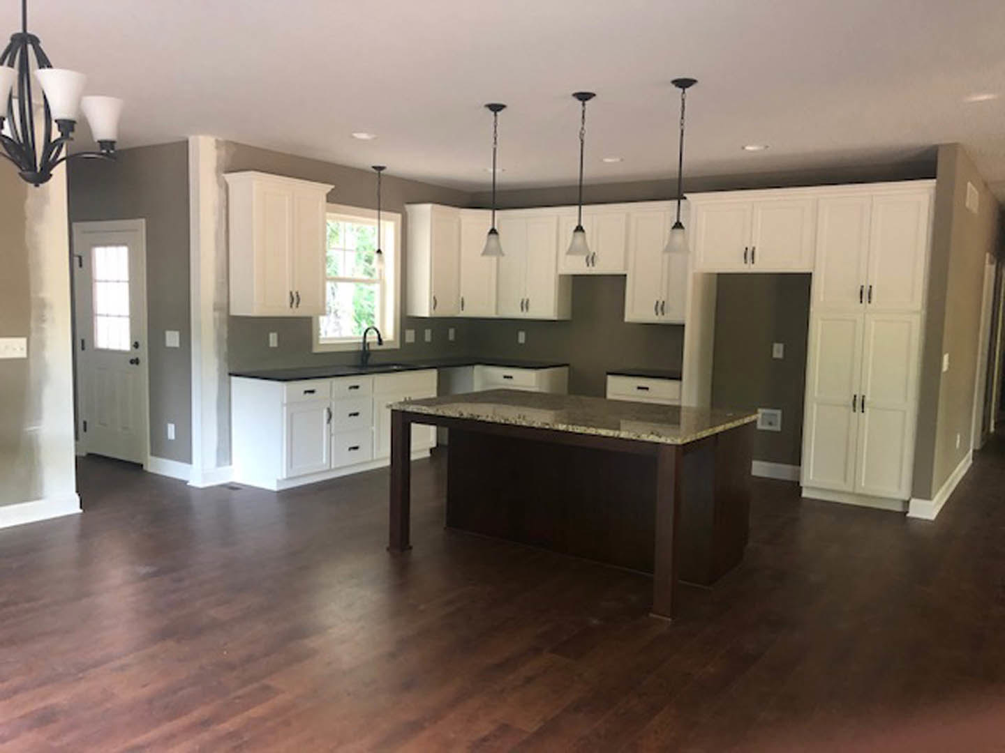 Open-concept kitchen featuring wood flooring, white cabinetry, stone countertop, central dining table, modern chandelier, and a windowed door allowing natural light.