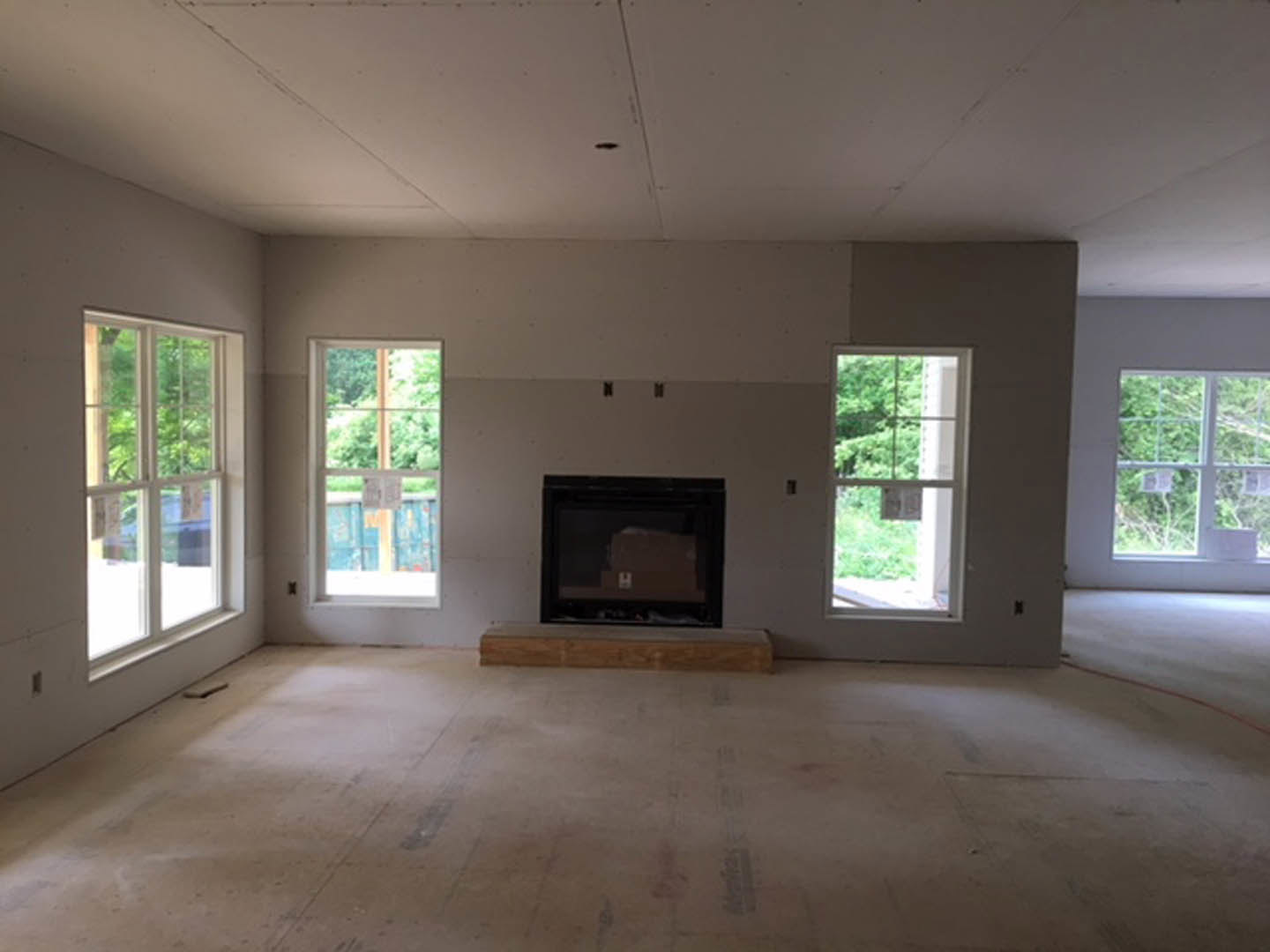 Living room with stone fireplace, large windows overlooking trees, hardwood floors, and white walls