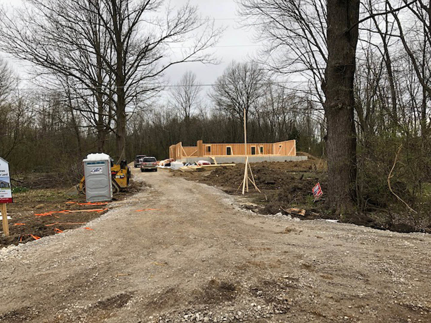 Dirt road lined with trees leading to a partially built structure with exposed framing and construction materials; grey shipping container and yellow caution sign visible nearby